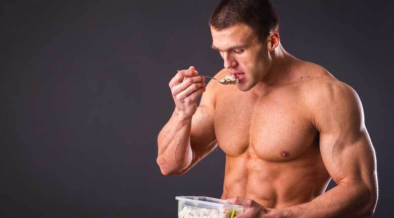 Подкаченный спортсмен кушает ложкой из пищевого контейнера сложные углеводы и белки